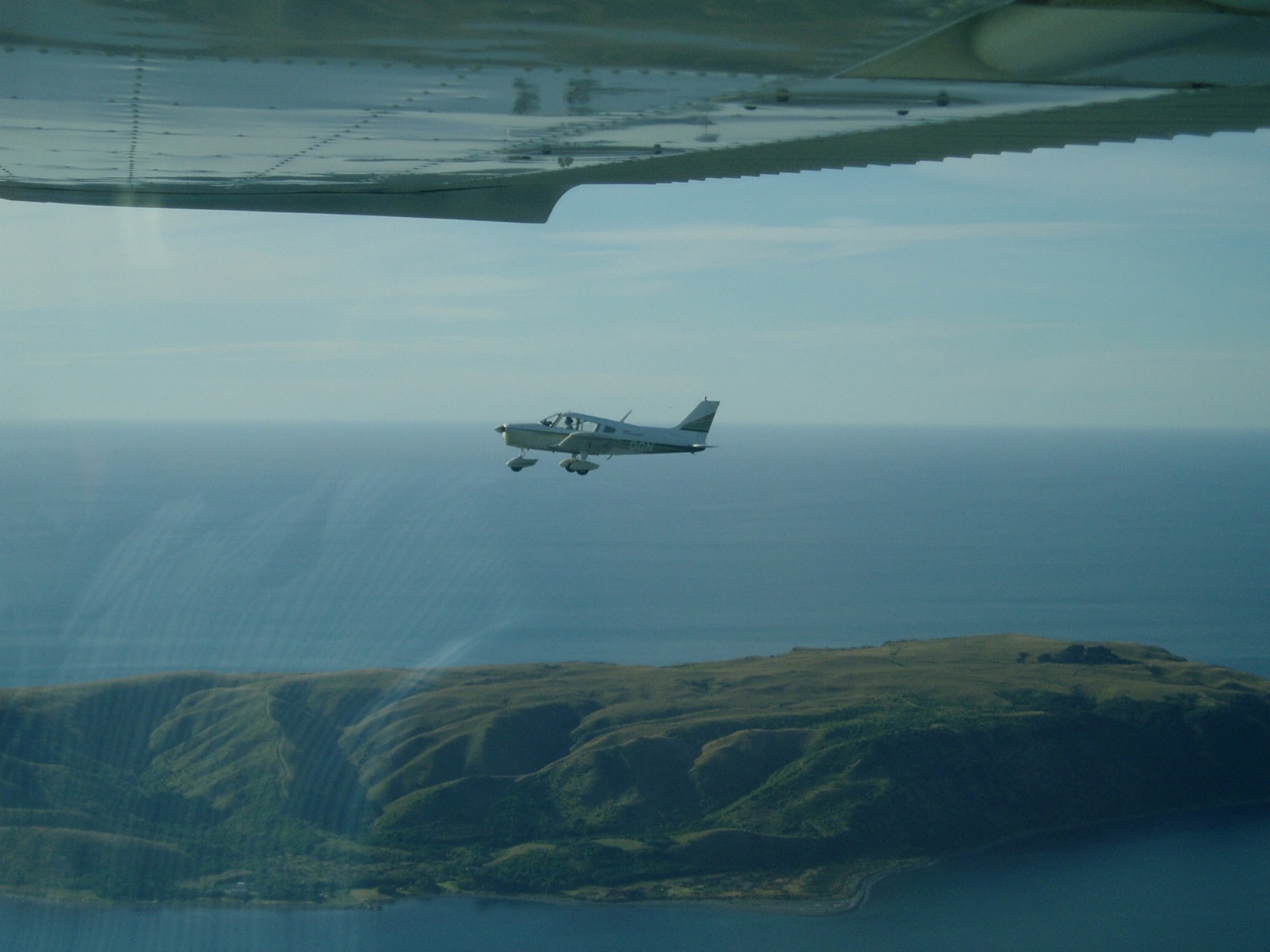 Light aircraft in flight over coastal hills, seen from beneath the wing of another aircraft
